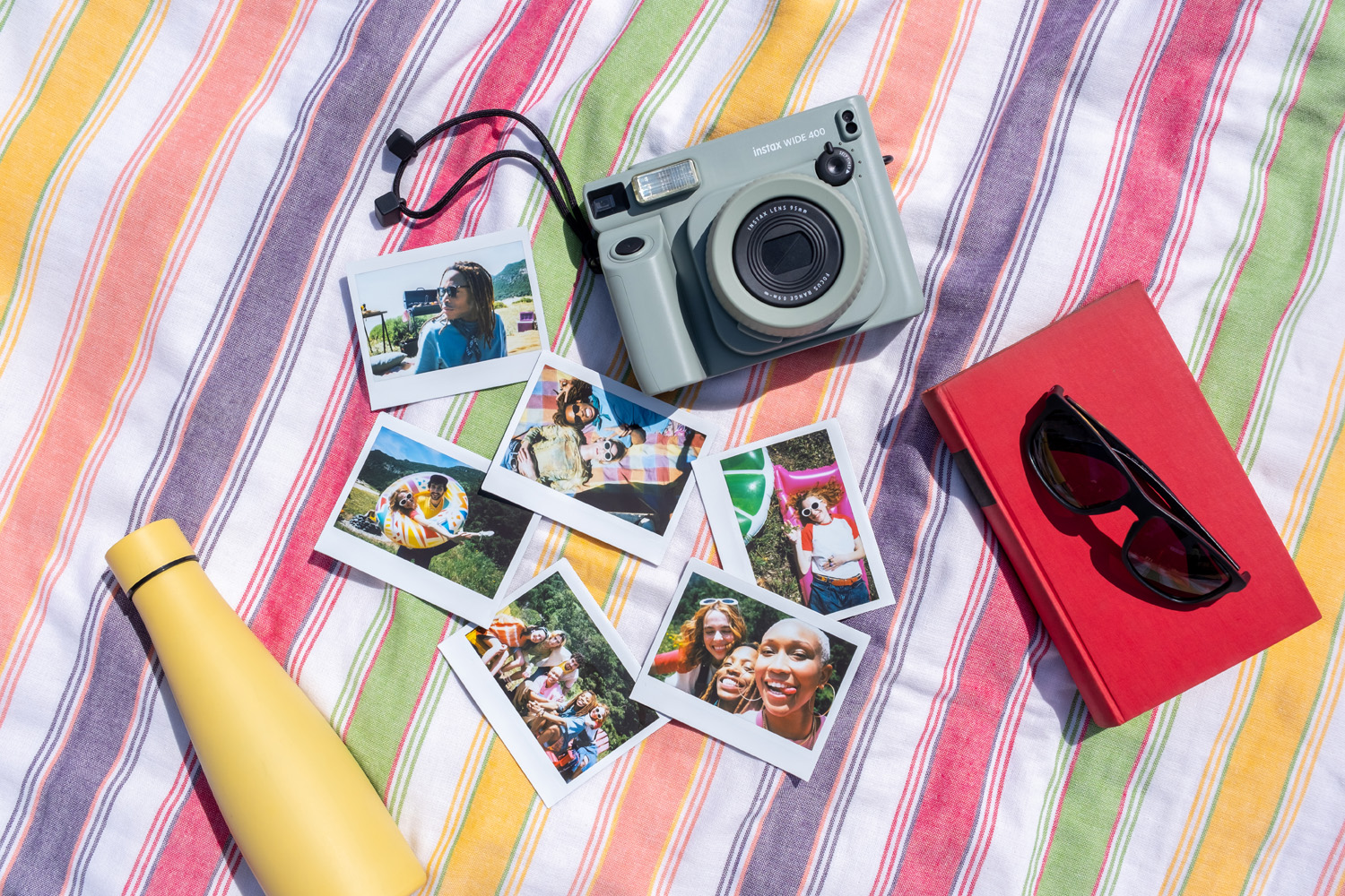 Плоская раскладка широкоугольной камеры Instax 400 и коллекция отпечатков на разноцветной полосатой скатерти.